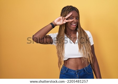 African american woman with braided hair standing over yellow background doing peace symbol with fingers over face, smiling cheerful showing victory 
