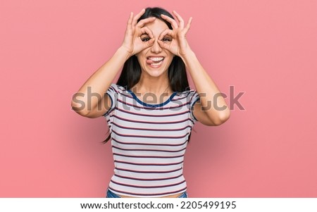 Young hispanic girl wearing casual striped t shirt doing ok gesture like binoculars sticking tongue out, eyes looking through fingers. crazy expression. 