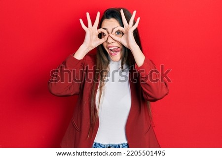 Young brunette teenager wearing business jacket doing ok gesture like binoculars sticking tongue out, eyes looking through fingers. crazy expression. 