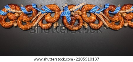 Oktoberfest concept - pretzels with traditional pattern ribbon and ears of wheat. A trendy hard light, dark shadow, black stone concrete background, banner format