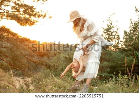 Portrait of laughing family wearing white dresses, walking in park forest illumined by sunset in summer. Young gorgeous woman mother holding upside down little girl daughter, playing. Relationship. Royalty-Free Stock Photo #2205515751
