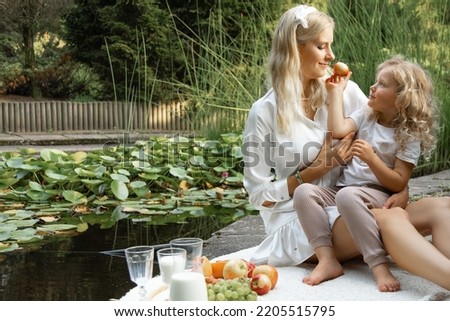 Portrait of wonderful family. Adorable little girl sitting with young woman on white plaid with fruits, jug of milk, offering apple to mother on edge of pond with green water lilies in summer. Picnic.