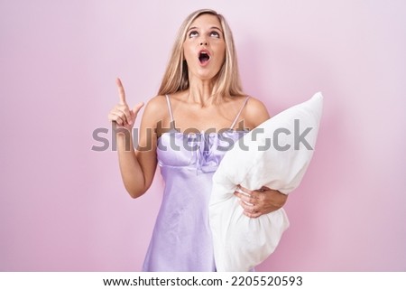 Young woman wearing pyjama hugging pillow amazed and surprised looking up and pointing with fingers and raised arms. 