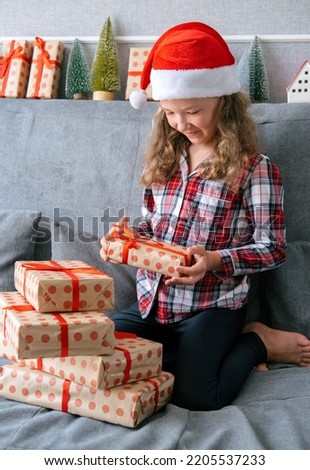 Cute candid portrait of little girl in Santa Claus hat and plaid woolen pajama with stack of present boxes.
