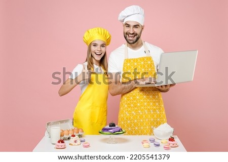 Teen girl dad father chef cook confectioner baker in yellow apron cap at table recipe on pc laptop show thumb up like gesture isolated on pink background Mousse cake food workshop master class process