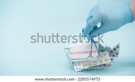 cropped view of person in latex glove holding shopping basket with coronavirus lettering on card and medicaments on blue