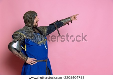 Side view portrait of man, medieval warrior or knight in chain armor pointing, giving instructions isolated over pink studio background. Leader. Comparison of eras, history, renaissance style Royalty-Free Stock Photo #2205604753