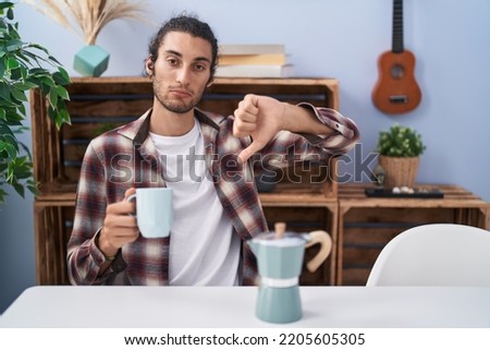 Young hispanic man drinking coffee from french coffee maker with angry face, negative sign showing dislike with thumbs down, rejection concept 