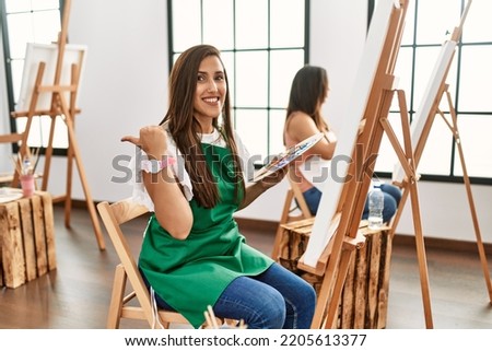Young hispanic artist women painting on canvas at art studio with a big smile on face, pointing with hand finger to the side looking at the camera. 