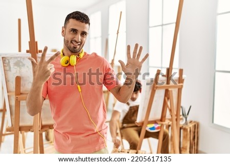 Young hispanic man at art studio showing and pointing up with fingers number eight while smiling confident and happy. 