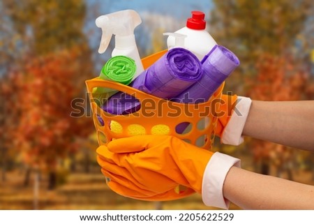 Woman's hands in rubber protective gloves holding orange basket with garbage bags, bottles of glass and tile cleaner, sponge with blurred autumn trees on the background. Washing and cleaning concept.