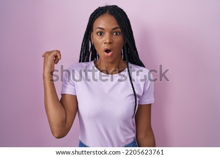 African american woman with braids standing over pink background surprised pointing with hand finger to the side, open mouth amazed expression. 