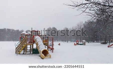 It is snowing in large flakes in the city. A blizzard is covering a playground with play structures on the edge of the lawn and surrounded by trees in the city park Royalty-Free Stock Photo #2205644551
