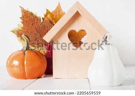 Autumn house insurance, sale, rent concept. White decorative pumpkins and autumn yellow leaves with miniature wooden house close-up and copy space..