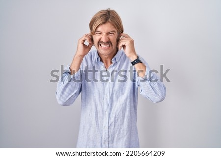 Caucasian man with mustache standing over white background covering ears with fingers with annoyed expression for the noise of loud music. deaf concept. 