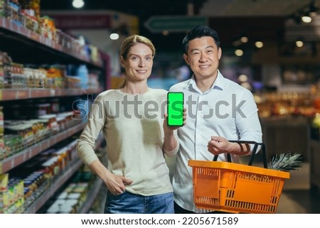 Young family diverse couple of shoppers in supermarket, smiling and looking at camera, grocery department, man and woman holding shopping basket and showing green screen of smartphone