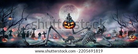 Halloween - Zombie Skeleton With Pumpkin In Hand Sitting On Cemetery At Night With Full Moon Royalty-Free Stock Photo #2205691819