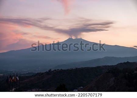 Smoke emitting from Mount Etna with orange sky in background. Beautiful tourist attraction with luxurious Elios hotel in foreground. Scenic view of dramatic landscape during sunset.