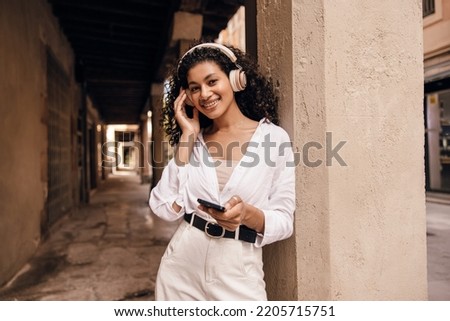 Positive young african girl uses her smartphone listening music with wireless headphones outside. Brunette with curly hair wears casual clothes. Concept wireless gadgets, use.