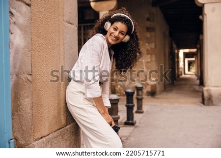 Funny young african woman with wireless headphones listens music outdoor in summer day. Girl with brunette hair wears shirt and white jeans. Concept modern technologies, good mood.