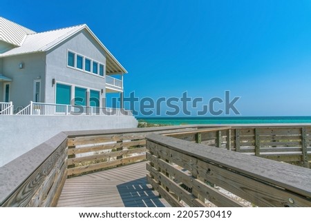 Beach house view from a boardwalk on a beach at Destin, Florida. Wooden boardwalk with a view of a beach house with veranda and picture windows against the sea and blue sky background.