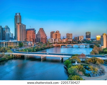Park and bridges over the Colorado River near the Austin, Texas cityscape during sunset. There are people at the park on the right and a viewof buildings with sunset glow against the sky horizon.