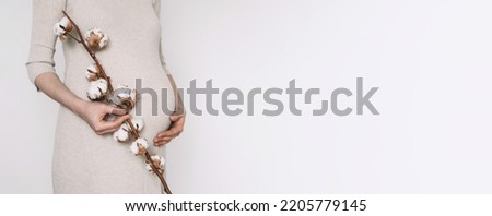 Pregnancy, maternity, preparation, baby expectation concept. 40 weeks of pregnancy. Pregnant woman in beige dress, touching belly, cotton flower on white background.