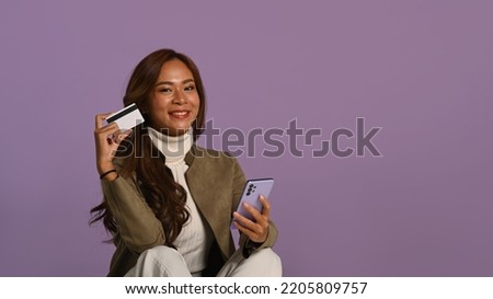 Studio photo of smiling young woman holding mobile phone and credit card sitting on purple background