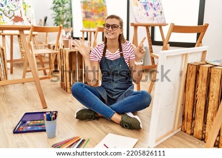 Young brunette woman at art studio sitting on the floor showing and pointing up with fingers number seven while smiling confident and happy. 