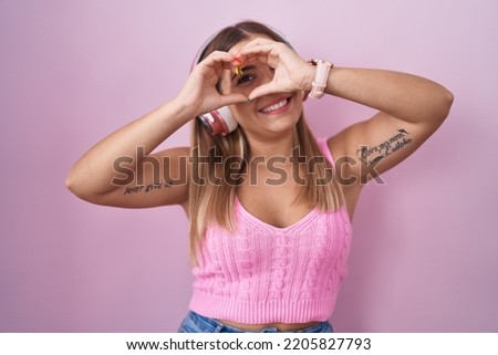 Young blonde woman listening to music using headphones doing heart shape with hand and fingers smiling looking through sign 