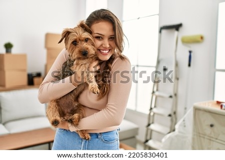Young beautiful hispanic woman smiling confident hugging dog at new home Royalty-Free Stock Photo #2205830401
