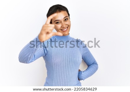 Young hispanic woman with short hair standing over isolated background smiling and confident gesturing with hand doing small size sign with fingers looking and the camera. measure concept. 