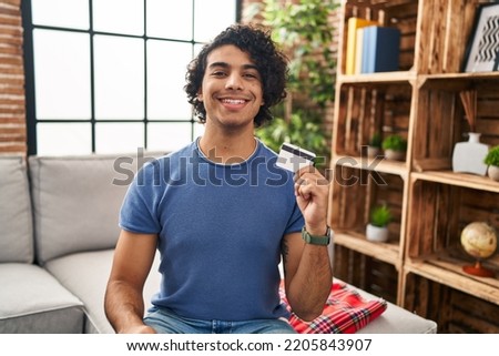 Hispanic man with curly hair holding credit card looking positive and happy standing and smiling with a confident smile showing teeth 