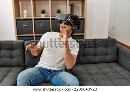 Young hispanic man scared playing with virtual reality glasses sitting on the sofa at home.
