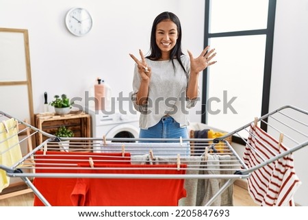 Young hispanic woman putting fresh laundry on clothesline showing and pointing up with fingers number seven while smiling confident and happy. 