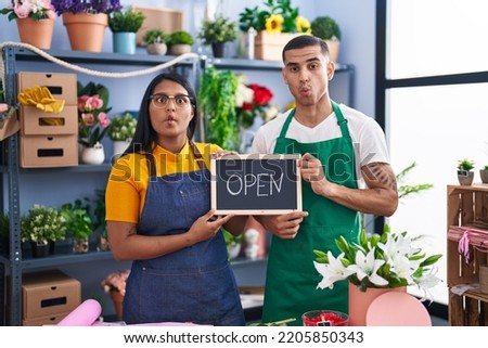 Young hispanic couple working at florist with open sign making fish face with mouth and squinting eyes, crazy and comical. 