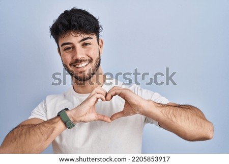 Hispanic man with beard standing over white background smiling in love doing heart symbol shape with hands. romantic concept. 
