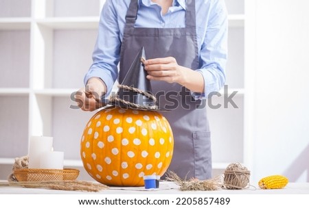 Woman in apron painting Halloween pumpkin, at wooden table, preparing holiday decorations at home
