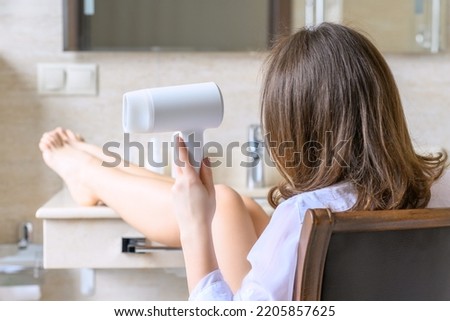 Back view of woman in a white nightgown blow dry hair by hair dryer sitting on armchair and looking in front of the mirror in the bathroom