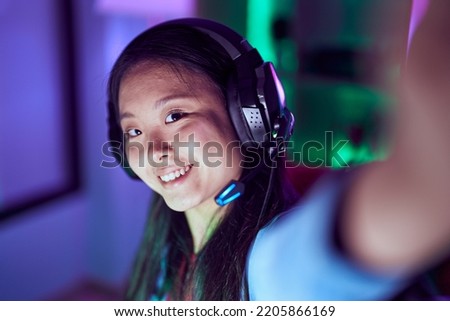 Young chinese woman streamer smiling confident make selfie by camera at gaming room