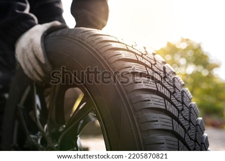 Close-up of hands with gloves rolling a new winter tyre  in bright sunlight ready for tyre change. Driving safety, road safety and car service concept. Royalty-Free Stock Photo #2205870821