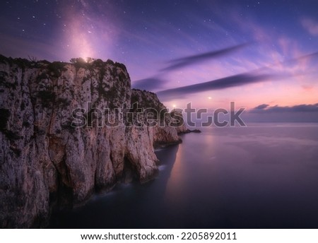 Lighthouse on the mountain peak at starry night in summer. Beautiful cliffs, rocky sea coast, bright stars, milky way and violet sky with clouds at dusk. Lighthouse of Cape Lefkada, Greece. Landscape Royalty-Free Stock Photo #2205892011