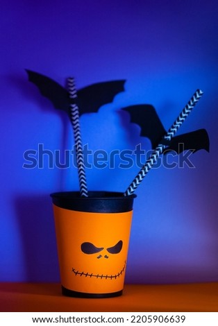 Halloween decor. Paper cup with straw inside and paper silhouettes of bats, bright background. Spooky holiday symbols