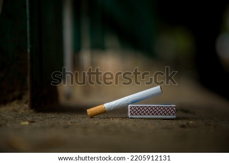 Cigarette and match box on the street , cigarette is standing by the support of matchbox.   