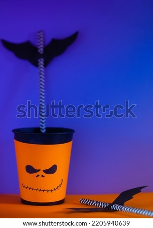 Halloween decor. Paper cup with straw inside and paper silhouettes of bats, bright background. Spooky holiday symbols, selective focus. Space for text