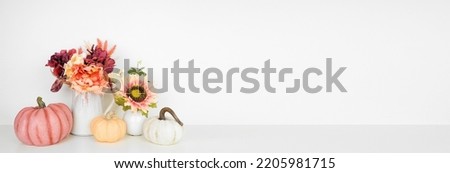Autumn decor on a white shelf against a white wall banner background. Pumpkins and flowers in vases with pink hue fall colors. Copy space.