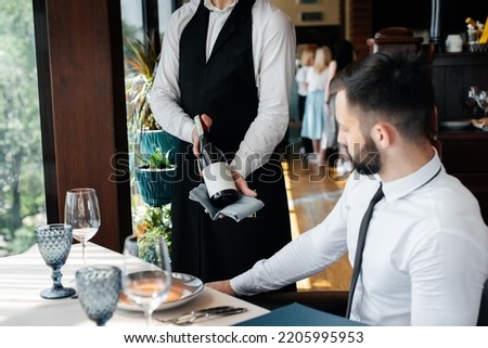 A young sommelier in a stylish uniform demonstrates and offers the client fine wine in the restaurant. Customer service. Table setting in a fine restaurant. Royalty-Free Stock Photo #2205995953