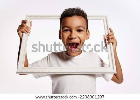 Wow, delight. Joyful happy african little boy in white tee stick out of picture frame and laughing isolated over white background. Kids emotions, happy childhood, facial expression, ad, beauty concept