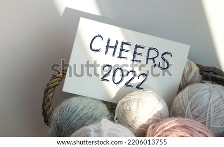 A card with the inscription CHEERS 2022 in a basket with balls of thread. Delicate colors, light background. Congratulations and wishes. Beginning of the year, beginning of spring.