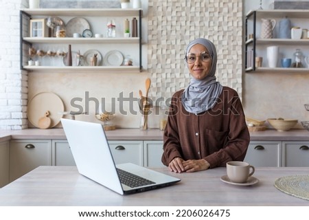 Portrait of young Muslim woman in hijab, woman in glasses looking at camera studying at home sitting in kitchen, female student using laptop for remote online learning